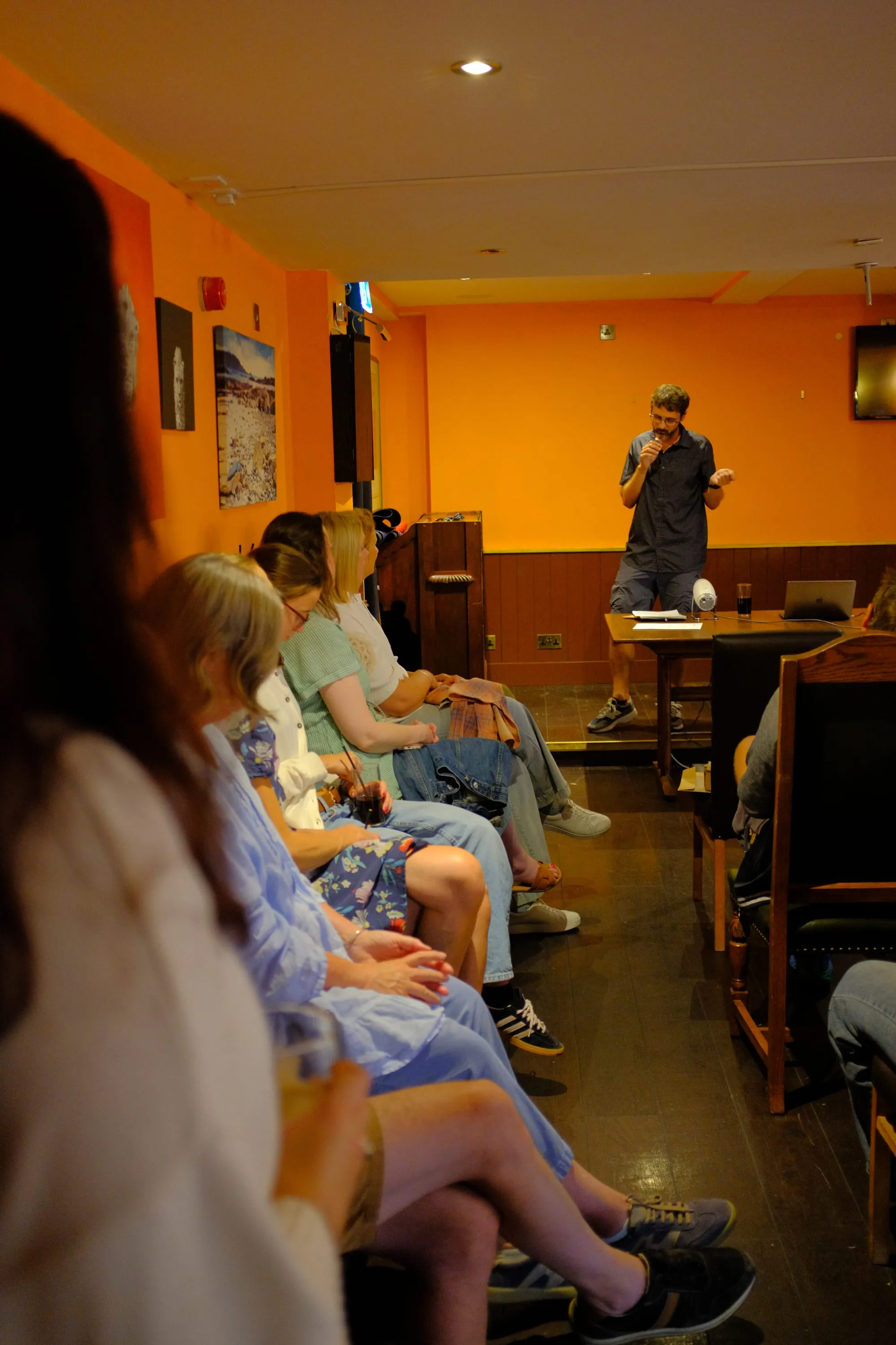 Nick Mann speaking into a microphone to a seated audience in the pub's back room