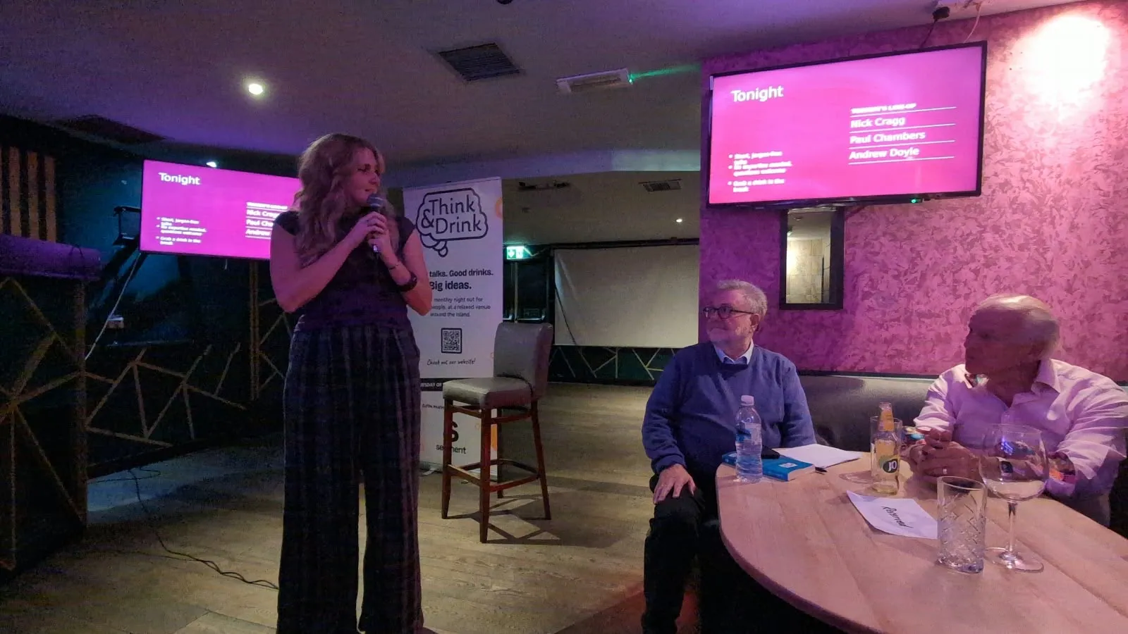 Sophie introducing the night with a microphone, the evening's line-up on the screen behind her